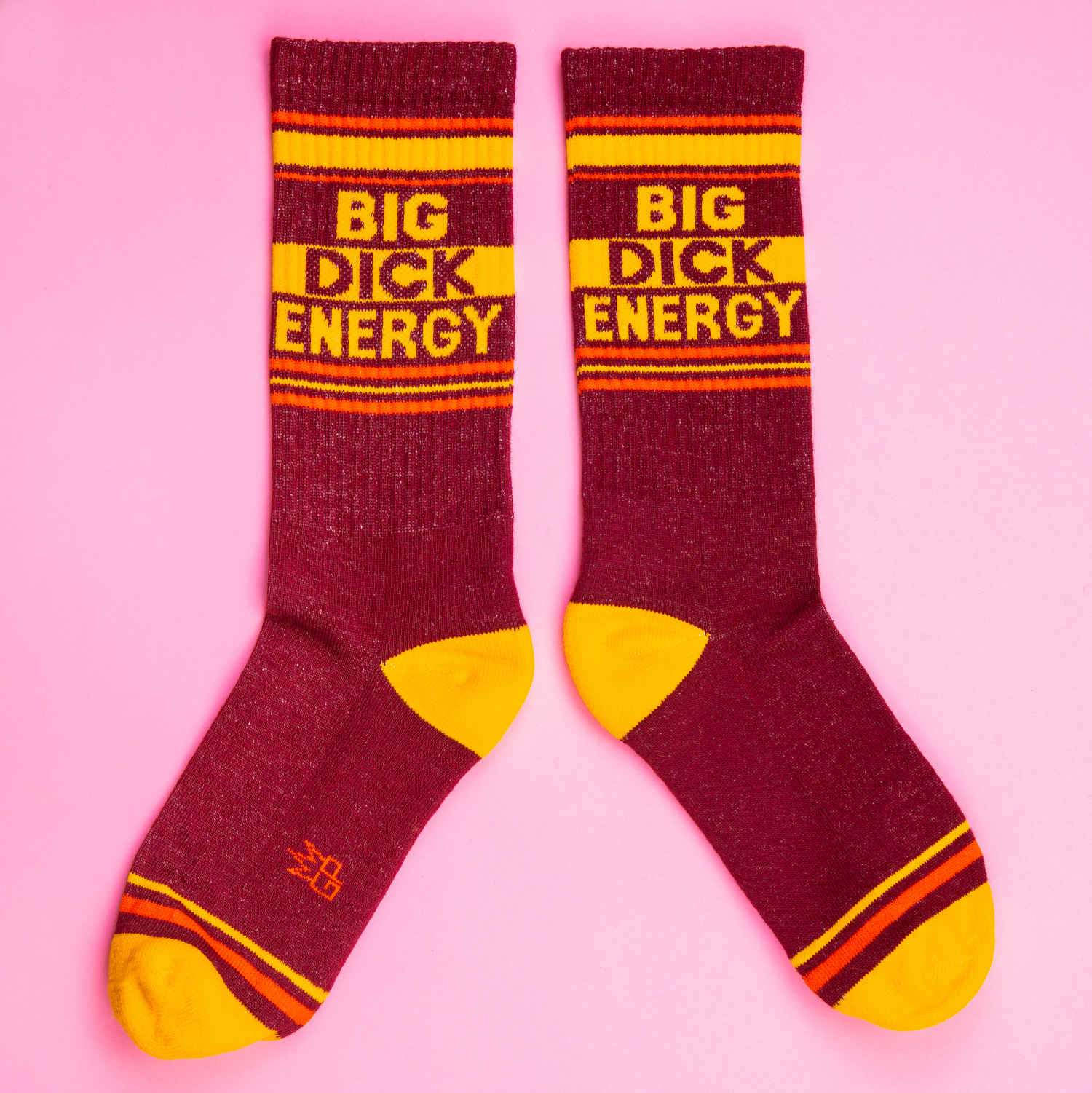 A pair of burgundy and yellow crew socks against a pink background that read "Big Dick Energy" with the Gumball Poodle logo along the arch.