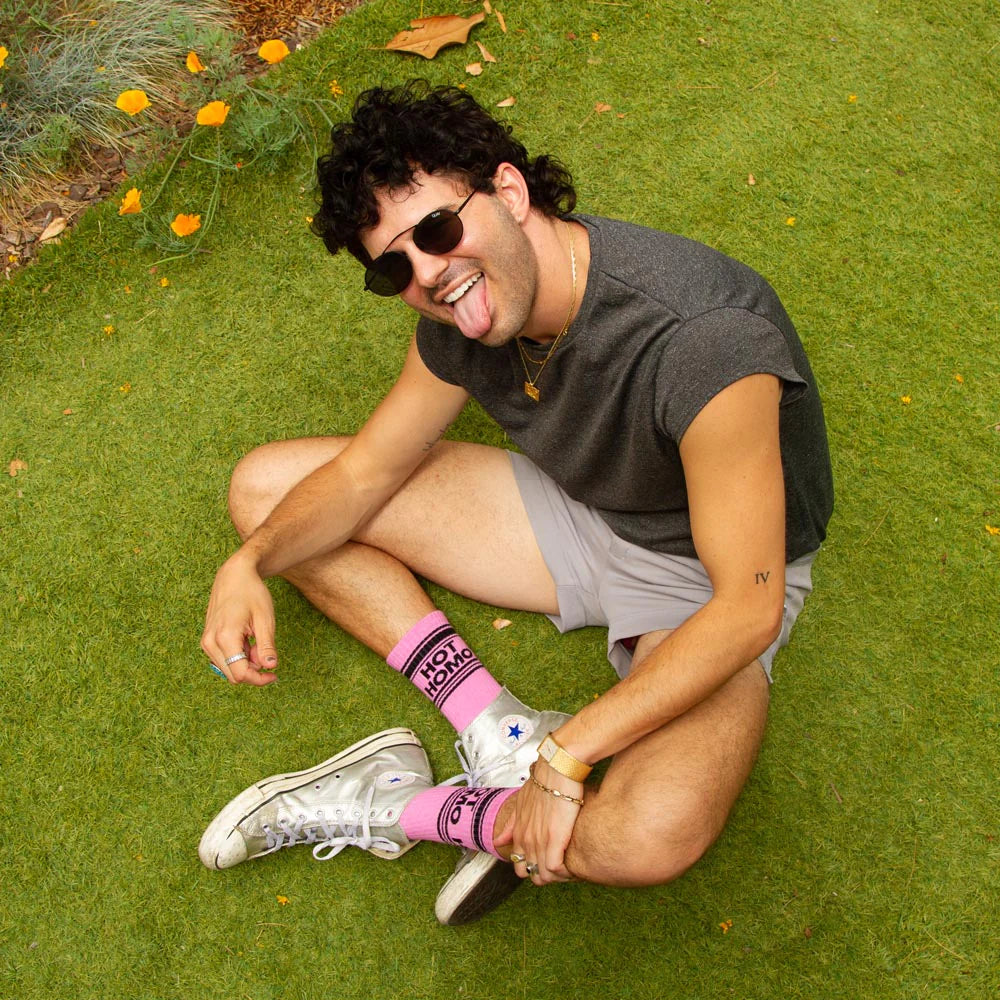 A man wearing a pair of hot pink and black crew socks that read "hot homo" demonstrated with silver Converse.